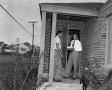Photograph: [Two men stand on the porch outside a house]