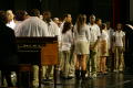Photograph: [Choir members stand on stage around mics]