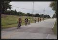 Photograph: [A line of cyclists biking down a road parallel to railroad tracks: L…