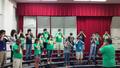 Photograph: [Students standing on a choral riser with their fingers on their nose…