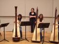 Photograph: [A row of three girls playing harps while a woman watches]