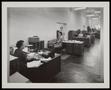 Photograph: [Women sitting at desks covered by papers in an office]