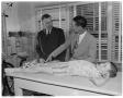Photograph: [Two men standing next to boy on examination table]