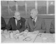 Photograph: [Two older men in suits, sitting at a table]