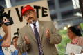 Photograph: [Hector Flores speaking at microphone wearing suit and red hat]
