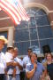Primary view of [Woman with a microphone addressing the crowd while a U.S. flag flies overhead]