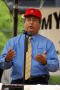 Photograph: [Man in red hat at microphone, gesturing with hands]