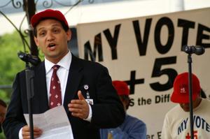 Primary view of object titled '[Man in suit and red hat speaking at microphone in front of printed sign]'.