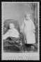Photograph: [A toddler sitting in a (tasseled) chair and a young child.]